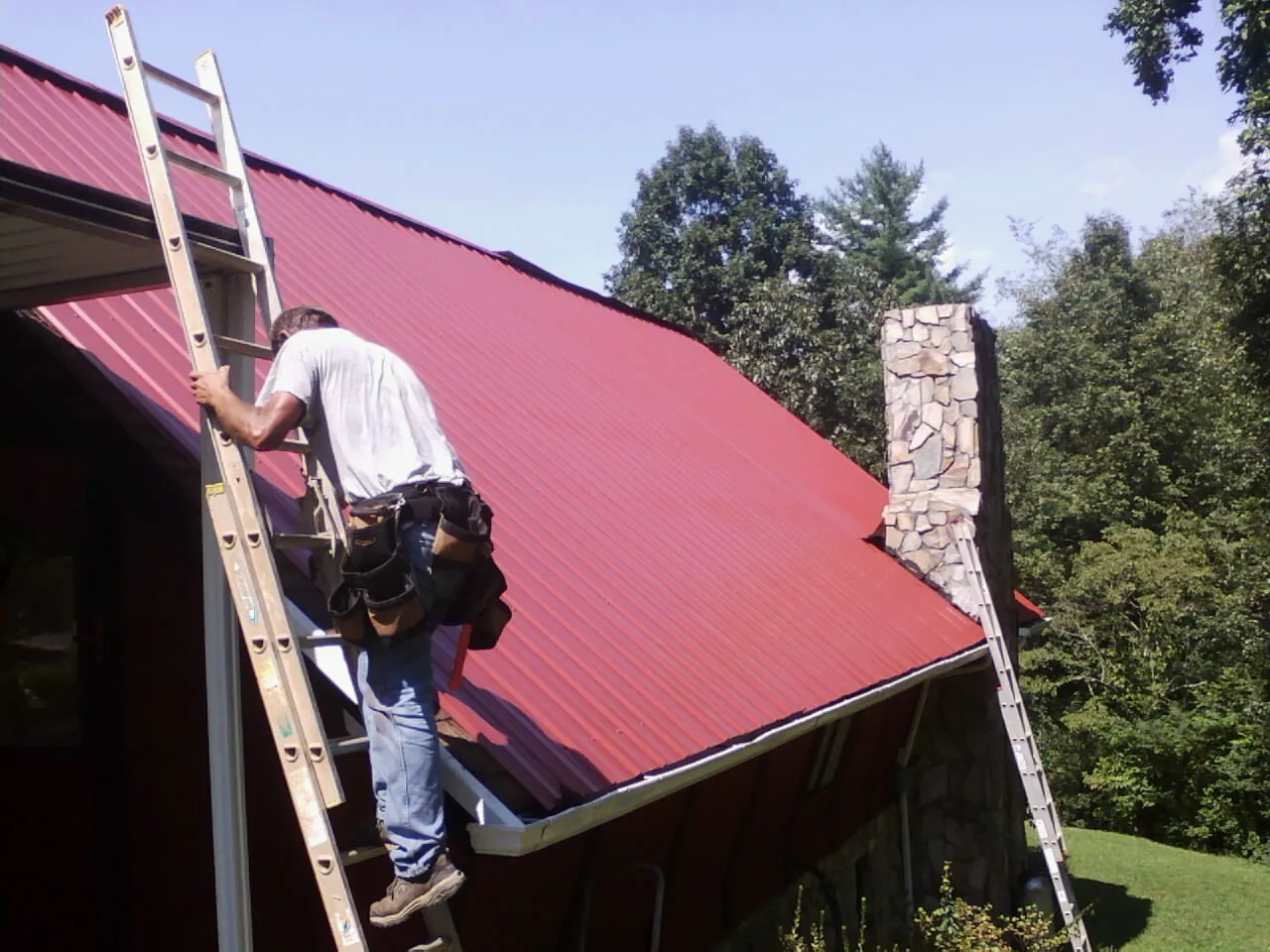 Roofer on a ladder beside a red metal roof and stone chimney — Soffit & Fascia Repair by Roofing Brothers in Schenley